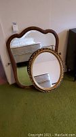 Two framed mirrors side by side, large wood framed with scalloped top and oval ornate framed mirror leaning against it.