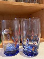 Photo showing four clear Murano glasses with colorful millefiori patterns and blue bases on a wood shelf.