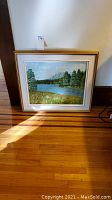Full view of framed oil painting showing a lake surrounded by pine trees and grassy foreground, in a gold-tone frame with white matting.