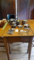 Wide view of the silver plated teapot, sugar, creamer, handled bowl, and assorted utensils spread on a wooden table.