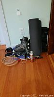 Photo showing a black rolled yoga mat, badminton rackets, Bauer inline roller skates, backpack and golf balls all placed on a wooden floor against a wall.
