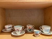 Five bone china tea cups on a wooden shelf, four of which have matching saucers underneath them. Floral patterns vary by cup.
