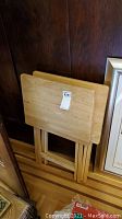 Photo of two wooden collapsible TV tray stands stacked and folded adjacent to a wall and framed artwork.