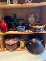 Shelves containing a collection of assorted pottery items including vases, bowls, mugs, containers, and pots in different colors and sizes, some with visible chips and a broken handle.