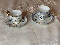 Photo showing two Reutter porcelain miniature coffee cups each with matching floral decorative saucers, placed on a light fabric surface.
