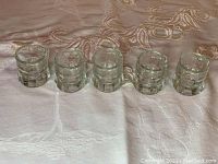 Five clear glass mini jug shot glasses in a row on a patterned fabric surface.