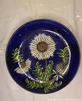 Top view of a round cobalt blue ceramic plate showing hand-painted traditional white flowers and green foliage.