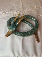 Full side view of the coiled green clay rattlesnake with duck beak head and golden antlers, laid on an embroidered fabric.