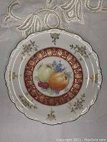Full top view of the ceramic plate showcasing the fruit painting in the center, gold decorative border, and floral designs on the rim.