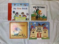 Top down view of four children's board books arranged flat on a fabric surface: 'My First Book,' 'Who Pretends?,' 'My Book of Opposites,' and 'Apes Find Shapes'.