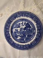 White ceramic plate with blue Japanese style design shown from the top, placed on fabric with embroidery.