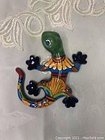 Top-down view of colorful ceramic lizard with a green head, blue hands and feet, and a multicolored body and tail, placed on a textured fabric background.