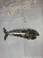 Fish-shaped decorative piece made from mother of pearl shell pieces on a fabric surface, showing top side with tail to left.