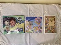 Three children's books laid out on a white cloth showing titles and illustrations 'Hush, Little One', 'Precious Nursery Rhymes', and 'The Mix Bedder Christmas'.