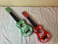 Pair of red and green ukuleles with white floral decoration on the bodies, lying on a white cloth.