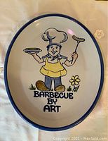 Top view of an oval stoneware serving plate with a chef carrying a sausage on a fork and a dish in the other hand, with 'Barbecue by Art' text and floral accents.