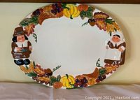 Front view showing the full oval plate with colorful raised relief border featuring a male Pilgrim on left, female Pilgrim on right, pumpkins, grapes, cornucopia and autumn leaves all around the border.