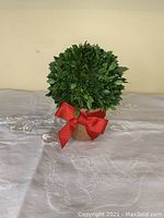Front view of the artificial topiary tree on a tan pot with red bow, sitting on a white lace tablecloth.