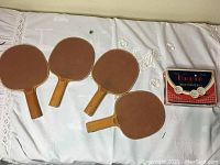 Four wooden ping pong paddles arranged side by side with a cardboard box of table tennis balls.