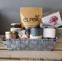 Gift basket items neatly arranged inside a gray woven basket including coffee bags, mugs, jam jar, candle, and other small gifts.