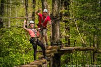 Two people on a wooden elevated platform among trees, wearing harnesses and helmets, enjoying zipline and aerial game trekking activities.