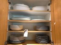 Image of cabinet shelves containing stacked dinner plates in two designs and three white ceramic serving dishes with lids on top shelf.