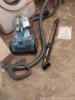 Full view of vacuum cleaner with hose, metal wand extension, and instruction manual on beige floor.