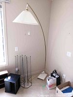 Photo shows full arched floor lamp next to window and corner wall with some other items around it. Lamp has brass-colored arched arm, white square base and white pleated shade.