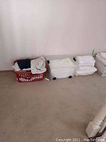 Wide view of laundry basket, plastic bin with linens, and stacks of folded linens on beige carpet near white wall.