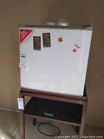 White Danby compact refrigerator shown standing on wooden stand, with a magnet and sticker on door, showcasing size and external condition.