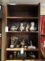 Wide view of bookshelf with multiple shelves holding various silverplate items including teapots, pitchers, sugar bowl, trays, and other servingware.