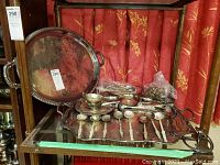 Silver plated serving trays, spoons, and spoon rest displayed on shelf with red curtain background, showing overall lot contents.
