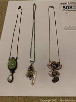 Three sterling silver necklaces laid flat showing pendants and chains including a green stone with teardrop shell, mixed stones with pearl, and hammered metal spiral pieces.