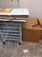 Photo showing white metal utility cart with flattened cardboard bankers boxes stacked on top and a large paper box beside it.
