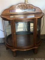 Front view of wood display cabinet with curved glass panels, decorative carved wood top with mirror inset, and wooden legs.