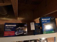 Photo showing Mastercraft 1/3 sheet sander box and Mastercraft 3/8 inch drill box next to drill bit set on a shelf in a basement.
