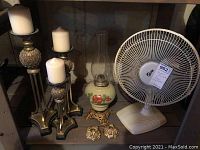 Photo of electric fan, candle holders with pillar candles, oil lamp, and two small decorative candle holders on a shelf in basement.