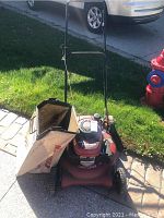 Side view of red Craftsman mulcher lawn mower with beige Dust Blocker bag attached, positioned on driveway beside grass and street curb