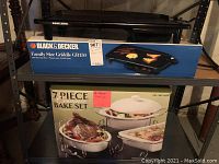 Two kitchen items on a metal shelving unit: Black & Decker Family Size Griddle GR150 in its original box and a 7-piece ceramic bake set also in its box. Griddle black with warming tray visible.