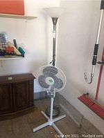 Full view showing both the torchiere floor lamp with metal base and the standing fan positioned in front of it.