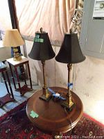 Pair of tall metal table lamps with black bell-shaped shades on round table, basement setting with unfinished walls and electrical panel in background.