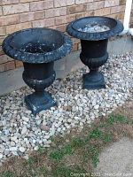 Pair of black resin urn-shaped planters placed outdoors on pebbled ground along a brick wall, showing overall view and relative size.