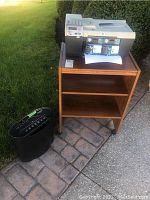 HP printer on top of brown MDF shelving unit with two shelves, two new packs of HP ink cartridges in original packaging in the printer's output tray, all placed outdoors on a stone driveway.
