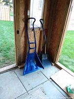Three snow shovels indoors near a window: two blue ergonomic shovels with double handles and one standard gray plastic shovel