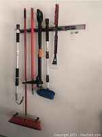 Overall view of brooms, brushes, tool, and metal wall bracket mounted on wall with tools hanging