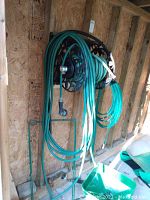 Two green garden hoses coiled on black metal hose winder mounted on wooden wall, hoses hanging down below
