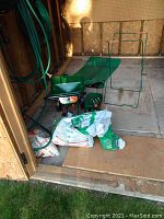 Photo showing Scotts Turf Builder Edge Guard mini seeder, Scotts hand seeder, part bags of peat moss and polymeric sand, all placed on plywood floor of a shed.