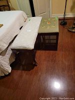 Two benches side by side, one white fabric upholstered with carved wooden legs, and one upholstered with vintage floral needlepoint.