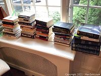 Wide view of 31 books stacked on a white radiator cover near a window, illustrating various sizes and titles mainly relating to classic literature, anthologies, and theatre.
