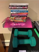 Two green 5-pound hand weights in front of a stack of assorted workout DVDs on a table with a pink Zumba Exhilarate box.